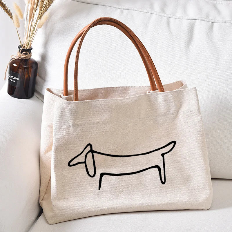 A beige cotton tote bag with a black dachshund print, featuring two brown leather handles, placed on a white sofa cushion next to a small glass vase with dried flowers.
