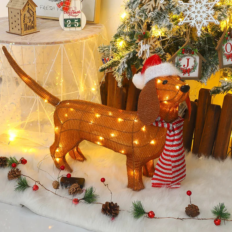 A light-up dachshund figurine with a red scarf, adorned with warm white LED lights, placed on a festive decorated floor near a Christmas tree.