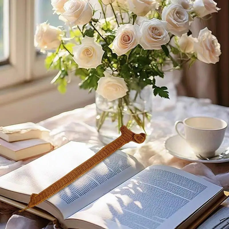 Open book with a bookmark, cup of tea, and vase of flowers on a table.